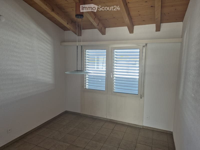 Empty room with exposed wooden ceiling beams, tiled floor, and two windows with blinds. A single hanging light fixture is visible.