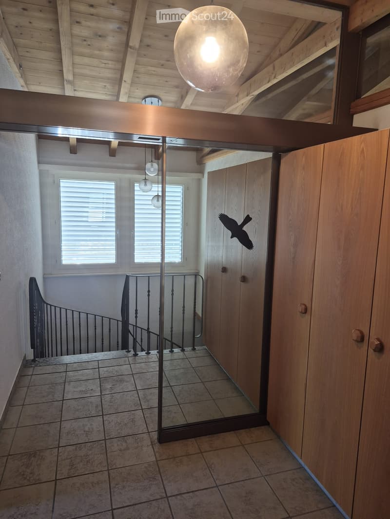 Modern hallway with wooden ceiling beams, pendant lights, tiled floor, and a staircase with a black metal railing. A large mirror reflects the space, and a decorative bird silhouette is visible on the wall.