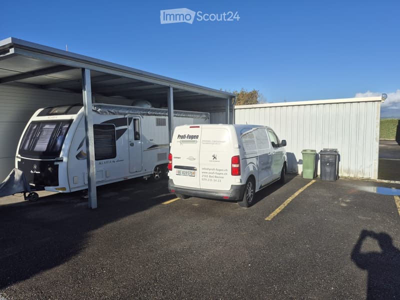 A caravan and a van are parked under a covered area with a white garage-like structure. The caravan is white with black accents, and the van has a white exterior with text and contact information.