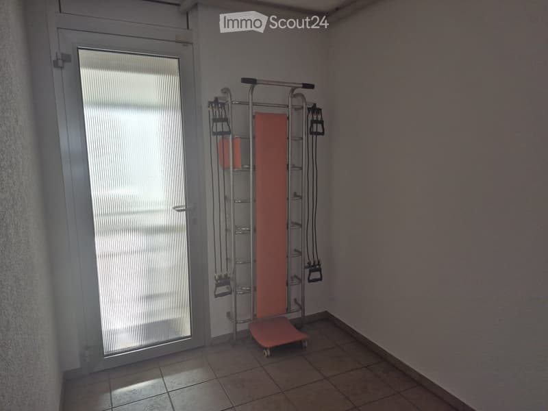 Empty room with tiled floor, white walls, and a large window with blinds. A small exercise bike is positioned near the window.