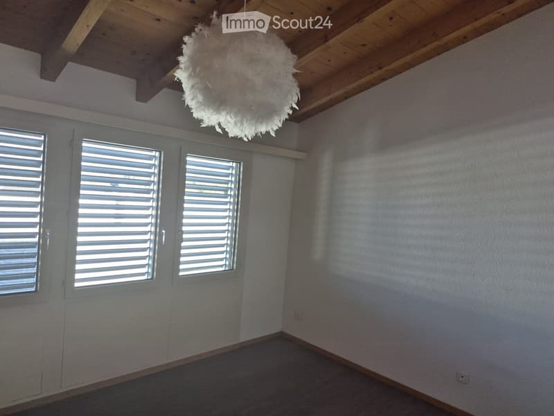 Empty room with white walls, wooden ceiling beams, and three windows with white shutters. A decorative white feather chandelier hangs from the ceiling.