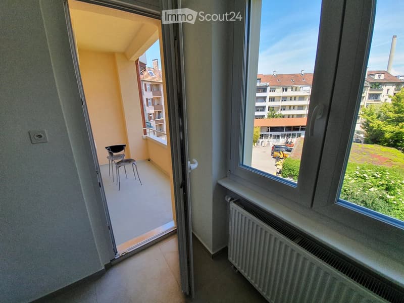 Open door to balcony, chairs, radiator, window with view of buildings and trees