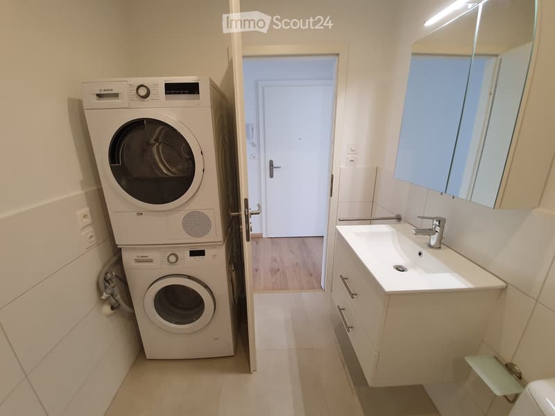 A bathroom with a white sink, mirror, and tiled floor. There is a white washing machine and dryer stacked on top of each other. The room has a white door and a wooden floor.
