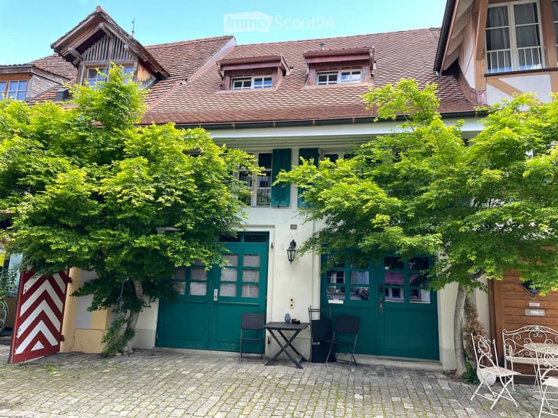 A charming garden area in front of a traditional house with a tiled roof, featuring a small seating area with a table and chairs, surrounded by lush green trees and a cobblestone pathway.