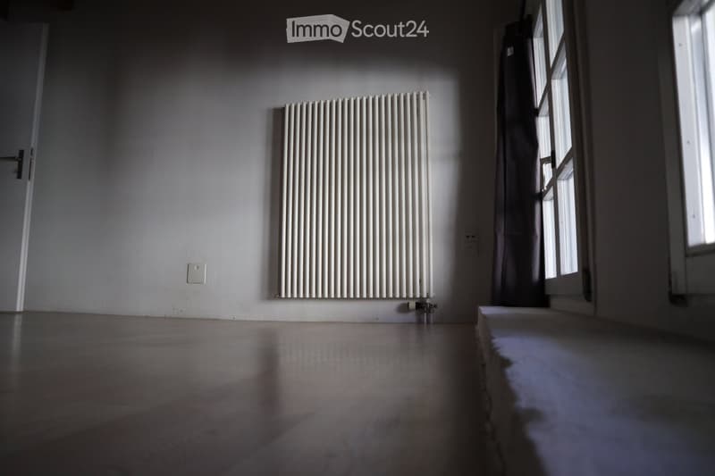 Empty room with wooden floor, white walls, radiator, and a window letting in natural light.