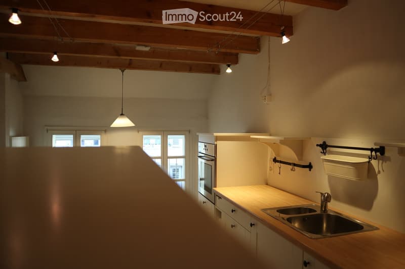 Bright kitchen with wooden beams on the ceiling, pendant lights, a stainless steel sink, and white cabinets.