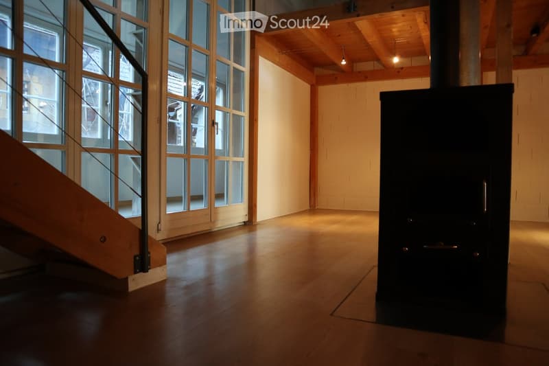 Empty room with wooden flooring, large windows, exposed wooden beams on the ceiling, and a black wood-burning stove.