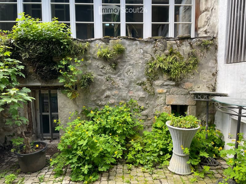 A small, well-maintained garden area with stone walls, potted plants, and a small table and chairs set against a white building with large windows.