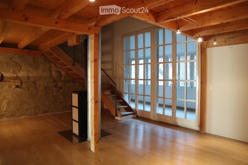 Empty room with wooden flooring, exposed wooden beams on the ceiling, large glass doors, and a wooden staircase leading upwards.