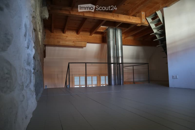 An empty room with wooden beams on the ceiling, a metal railing, and a stone wall. The floor is made of dark wood planks, and there is a large window in the background.