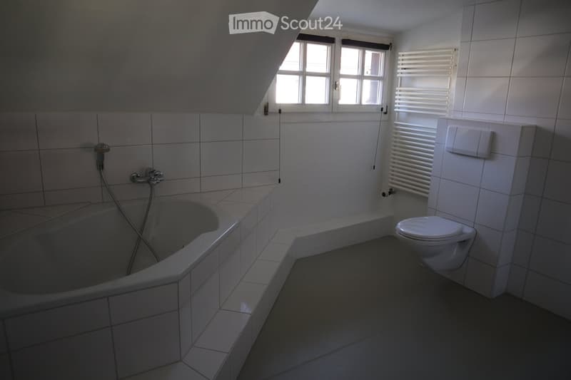 Modern bathroom with a bathtub, toilet, tiled walls, and a window with blinds. Includes a radiator and a faucet above the bathtub.