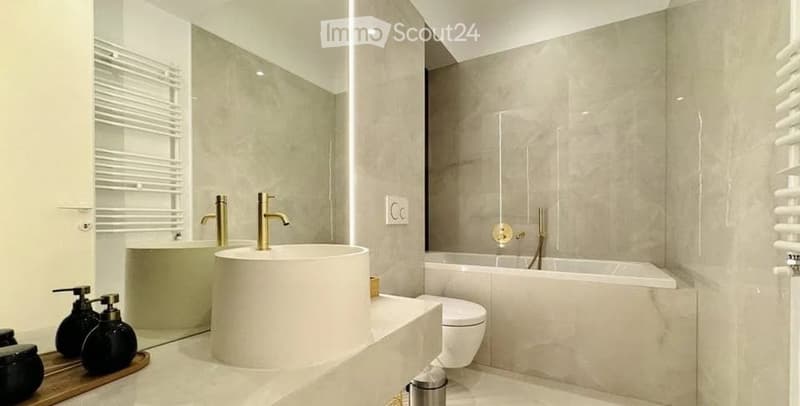 Modern bathroom with a large, round sink, sleek faucets, a bathtub, and a toilet. The space features light-colored tiles and a minimalist design.