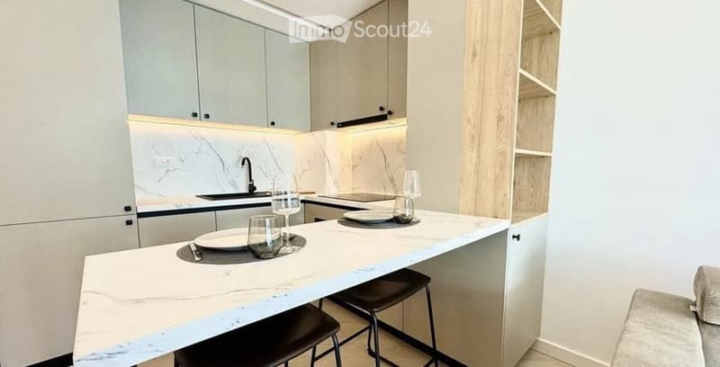 Modern kitchen with white cabinets, marble countertops, and built-in lighting. Includes a central island with stools, decorative glassware, and dishes.