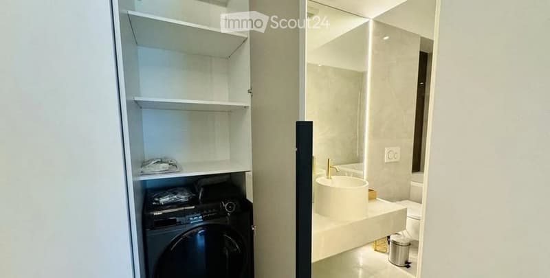 Modern bathroom with a white sink, gold faucet, large mirror, toilet, and a washing machine in a closet with shelves.