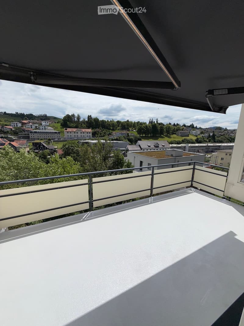 Balcony with white floor and modern railing, overlooking a town with houses, trees, and hills under a partly cloudy sky.