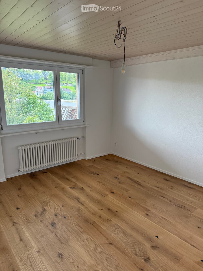 Empty room with wooden flooring, white walls, large windows, and a radiator. Exposed wooden ceiling beams and a single light fixture are visible.