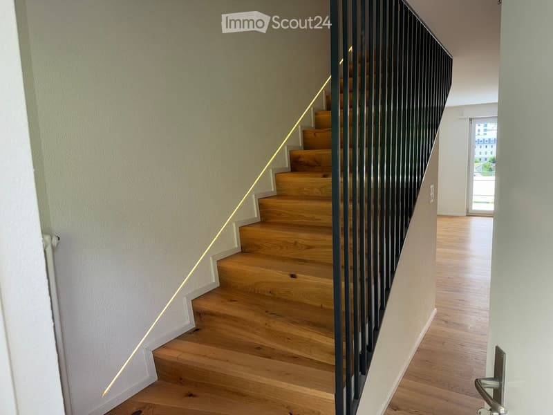 Modern hallway with wooden flooring, a staircase with wooden steps and a black metal railing, and a door leading to another room.