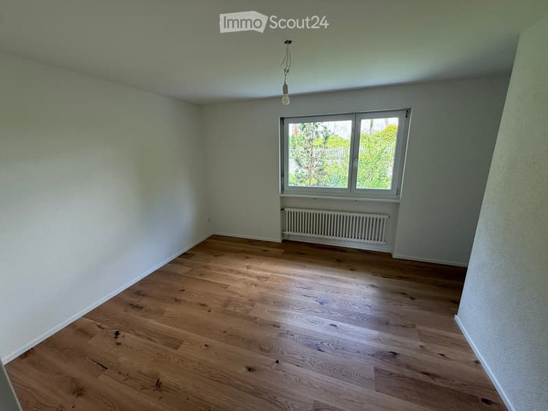 Empty room with wooden flooring, light green walls, a window with a view of greenery, and a radiator below the window.
