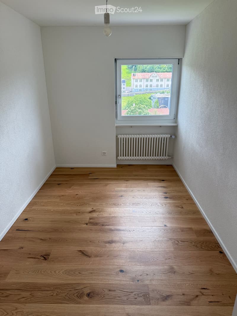 Empty room with wooden flooring, white walls, a window with a radiator below, and a hanging light fixture.