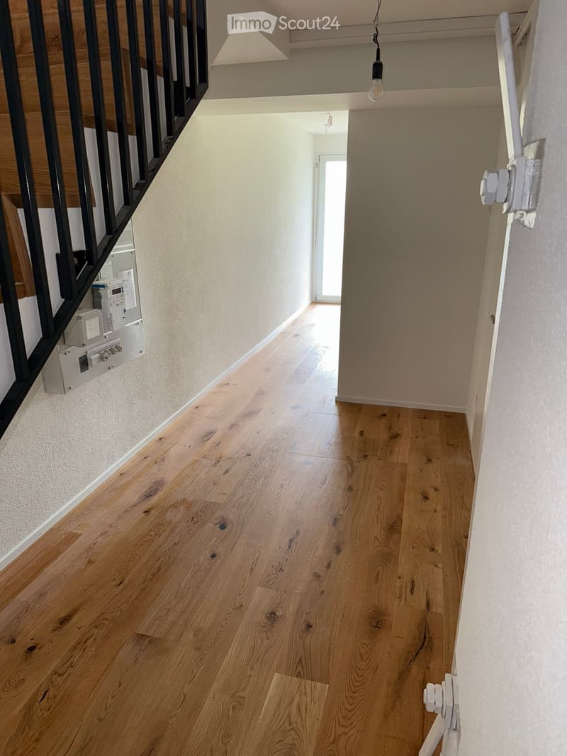 A narrow hallway with light wooden flooring, white walls, and a staircase with black railings visible on the left side. Natural light streams in from an open doorway at the end of the hallway.