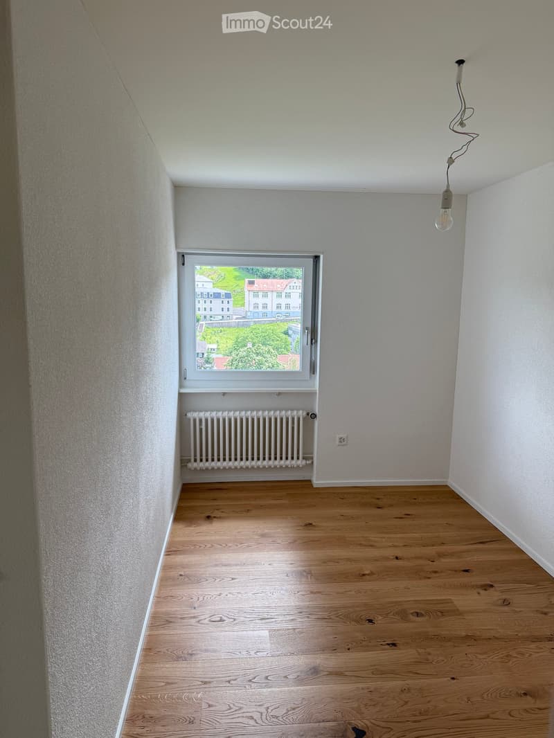 Empty room with wooden flooring, white walls, a window with a view of greenery, a radiator, and a single light fixture with a bulb.
