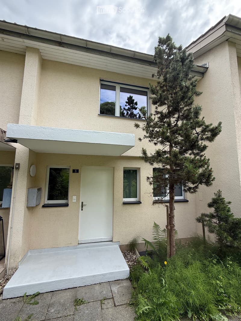 Two-story house with light beige exterior, white door, multiple windows, small garden with a tree and shrubs.