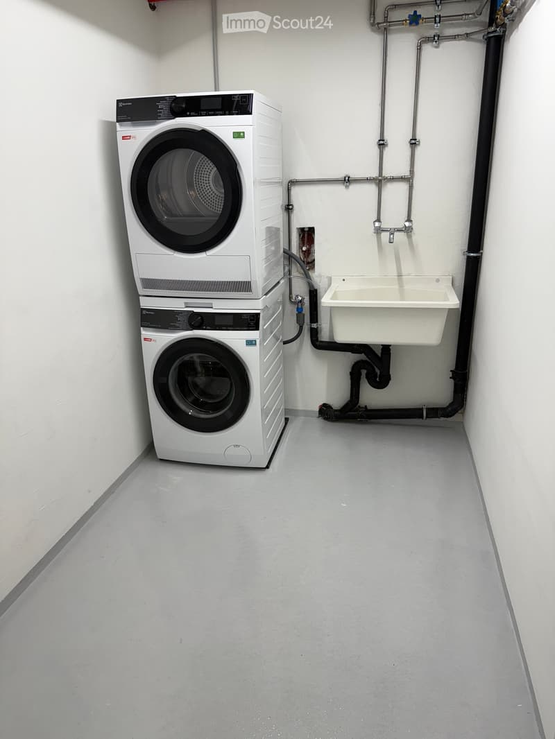 Compact room with a stacked washing machine and dryer unit, a small sink, and visible plumbing.