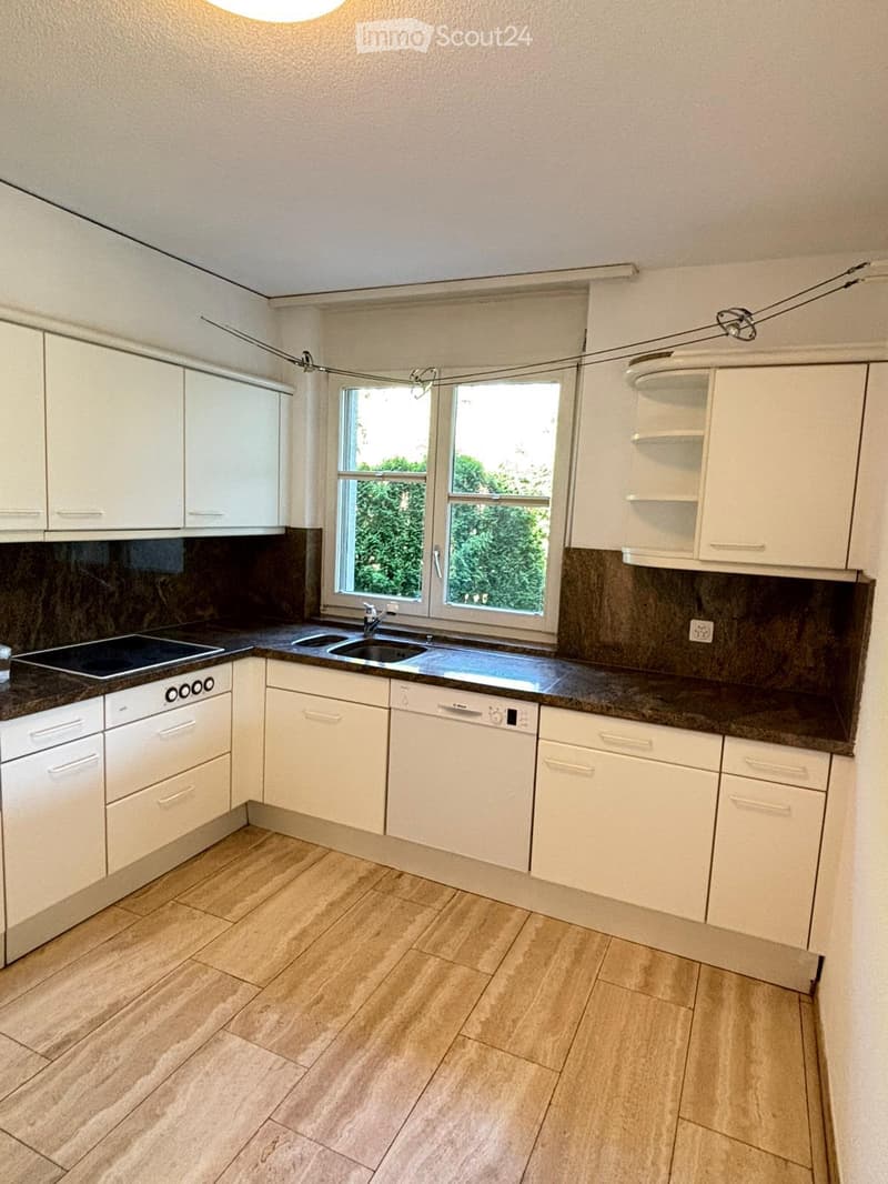 Modern kitchen with white cabinets, dark granite countertops, a sink, dishwasher, and electric stove. The room features wooden flooring and large windows with a view of greenery.