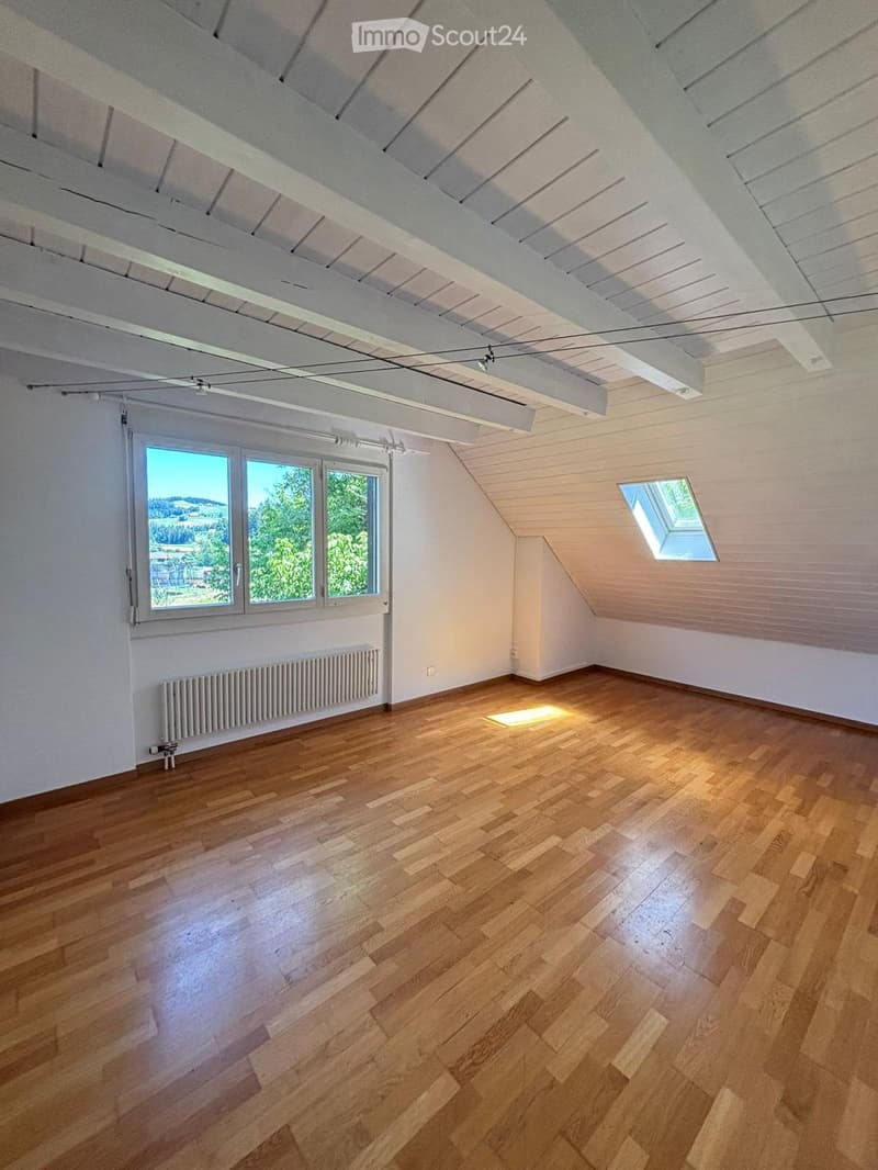 Empty attic room with wooden flooring, white walls, large windows, skylight, and exposed wooden beams on the ceiling.