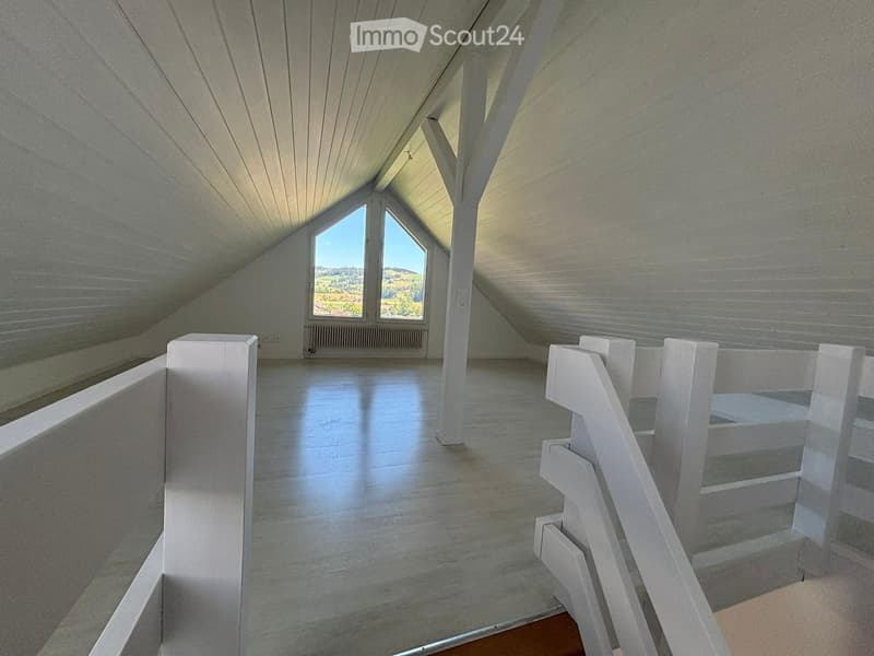 Bright, empty attic space with wooden floors, white walls, and large windows offering a scenic view. Features a staircase leading upwards.