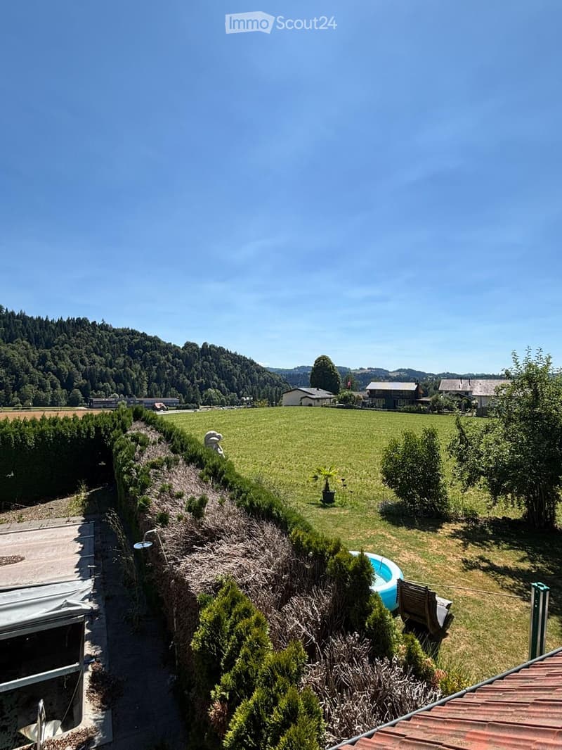A spacious garden with a swimming pool, surrounded by greenery and trees, with a scenic view of a valley and hills in the background.