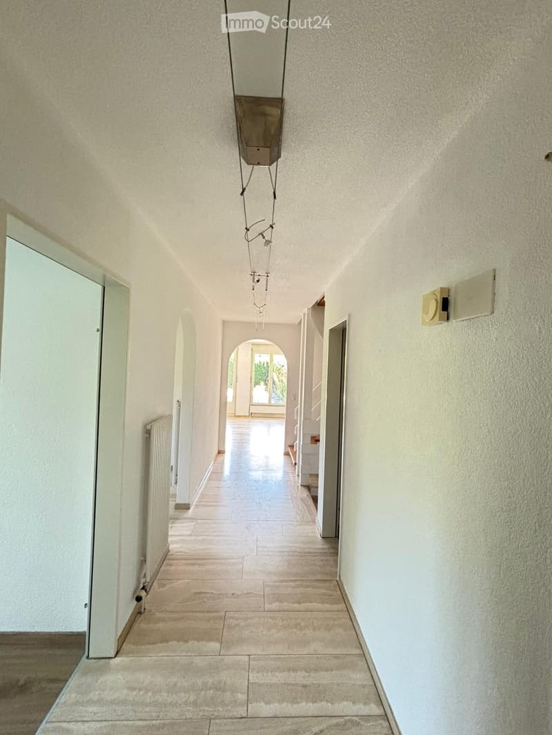 A long, narrow hallway with beige tiled flooring, white walls, and a modern hanging light fixture. The hallway features arched doorways leading to other rooms and a visible radiator on the left wall.