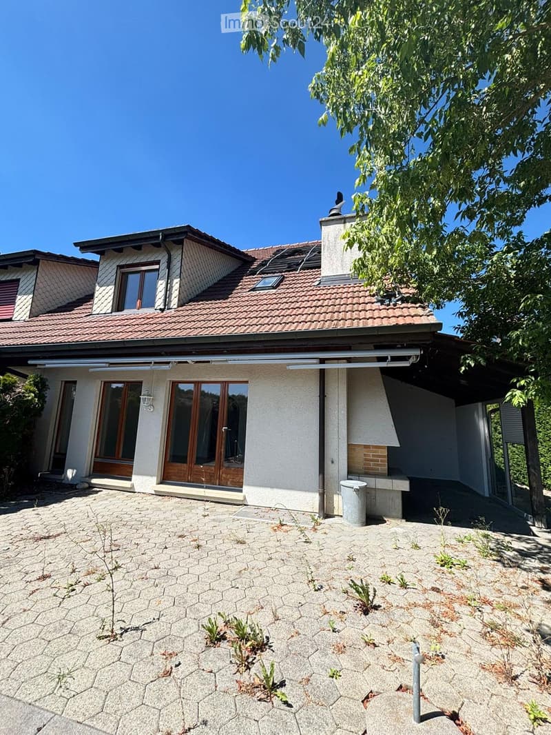 Two-story house with a tiled roof, white exterior walls, large windows, and a covered entrance area. The ground is paved with hexagonal stone tiles, and there is a small garden area with sparse vegetation.
