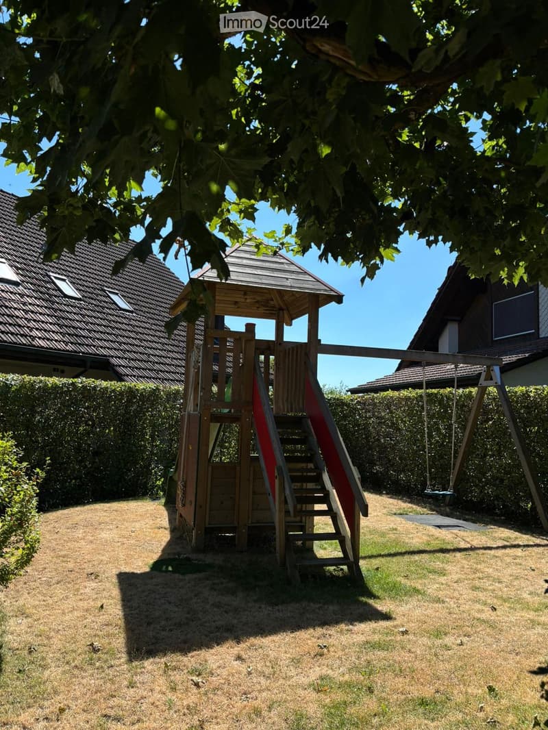 A well-maintained garden with a wooden playhouse, slide, swing, and surrounding houses.