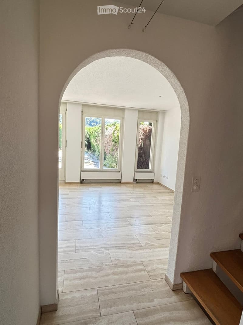 Empty hallway with beige walls, marble tiled floor, and an arched doorway leading to a bright room with large windows and radiators.