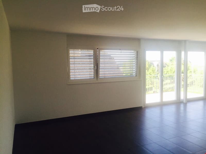 Empty room with dark tiled floor, white walls, large windows with blinds, and sliding glass doors leading to a balcony.