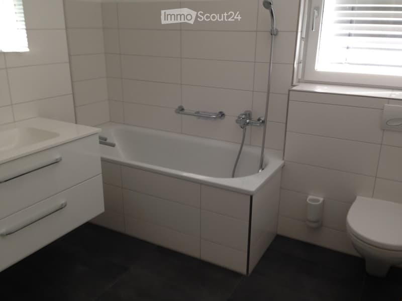 Modern bathroom with a bathtub, sink, toilet, and a handrail for safety. The walls are tiled, and the floor is dark.