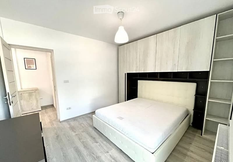 Modern bedroom with a white bed, built-in wardrobe, and shelves. The room features light wooden flooring and white walls.