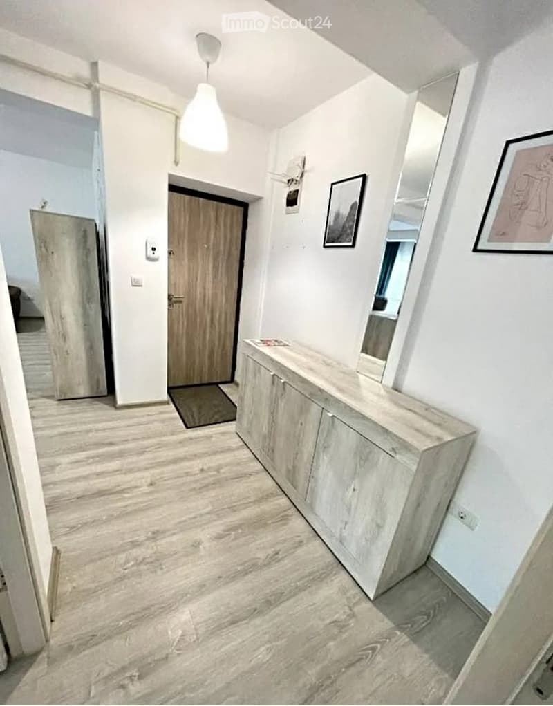 Modern hallway with light wooden flooring, white walls, a pendant light fixture, a wooden door, a sideboard with drawers, and framed artwork on the wall.