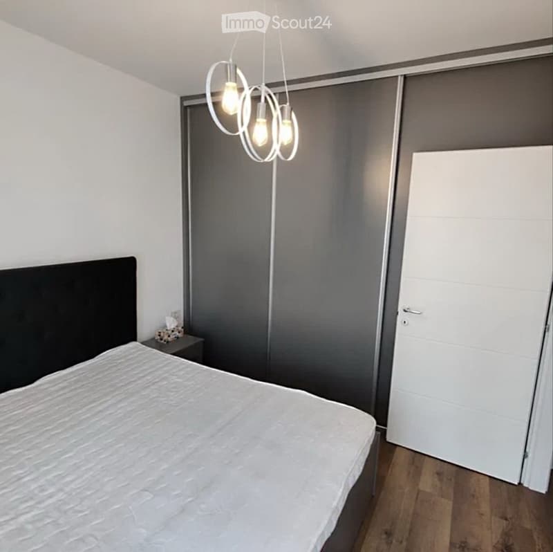 Modern bedroom with a large bed, nightstand, and built-in wardrobe. The room features wooden flooring and a contemporary pendant light fixture.