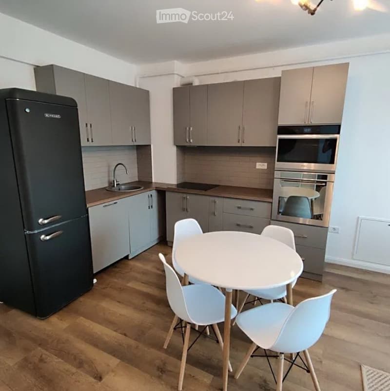 Modern kitchen with grey cabinets, stainless steel appliances, a round white dining table with four light blue chairs, and wooden flooring.