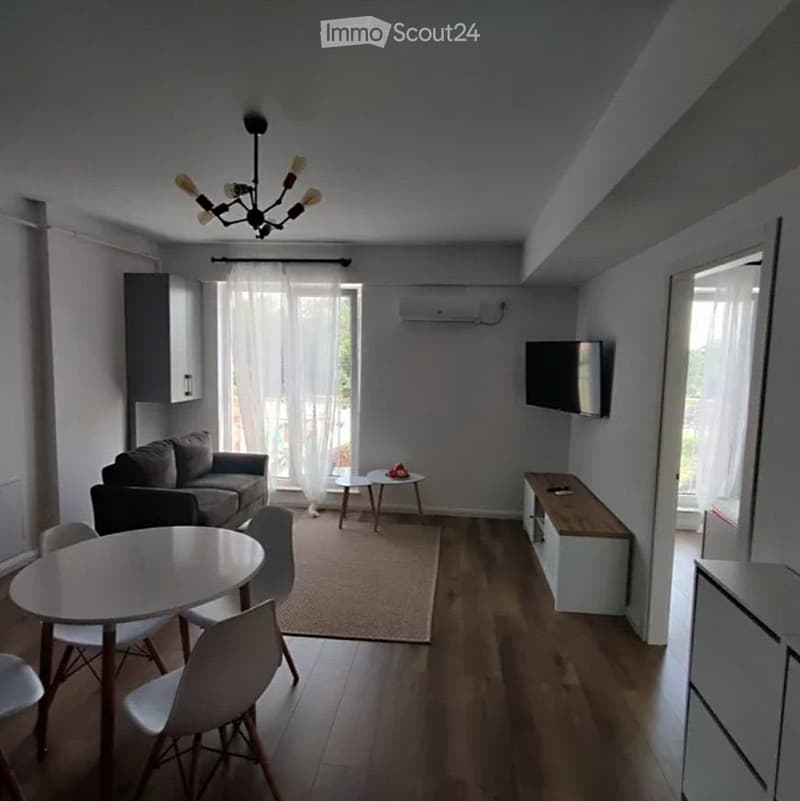 Modern living room with a sofa, dining table, chairs, TV, and a chandelier. The room features wooden flooring, large windows with sheer curtains, and a small rug in the center.