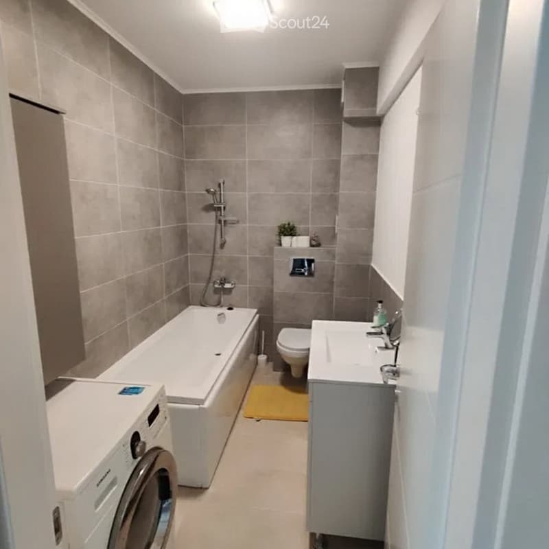 Modern bathroom with a bathtub, toilet, sink, and washing machine. Features grey tiled walls, a yellow bath mat, and a small plant on the shelf.