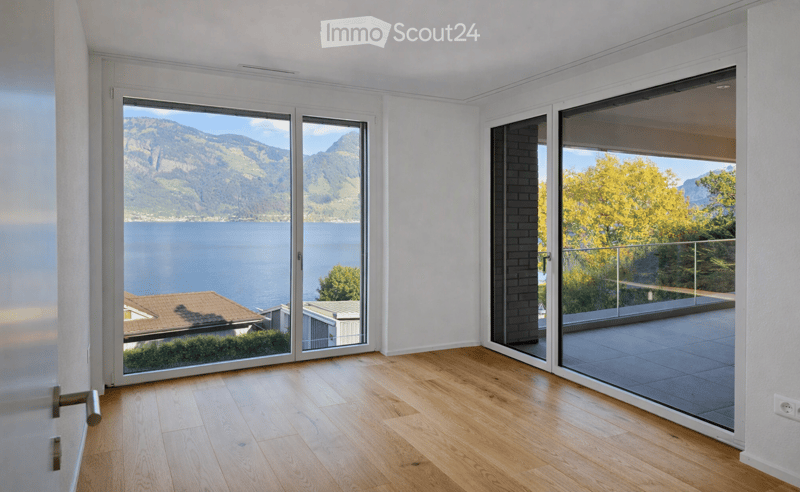 Bright, unfurnished room with large sliding glass doors offering scenic views of a lake and mountains. The room features light wooden flooring and white walls.
