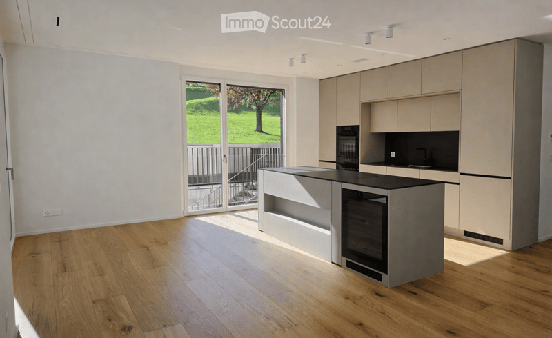 Modern kitchen with light wood flooring, white walls, and a central island with built-in oven. Large windows offer a view of a green outdoor area.