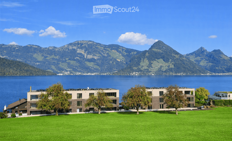 Modern residential buildings with large windows and balconies, surrounded by a well-maintained lawn and trees, with a scenic lake and mountain backdrop.