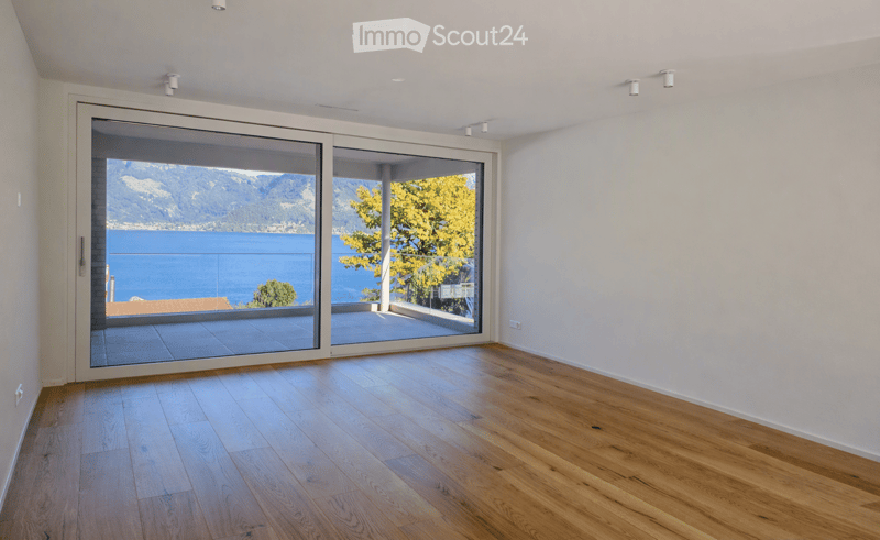 Bright, empty room with wooden flooring and large sliding glass doors opening to a balcony overlooking a scenic lake and mountains.