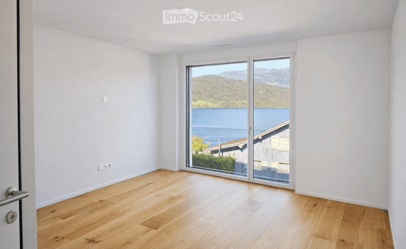 Empty room with light wooden flooring, white walls, and large sliding glass doors offering a scenic view of a lake and mountains.