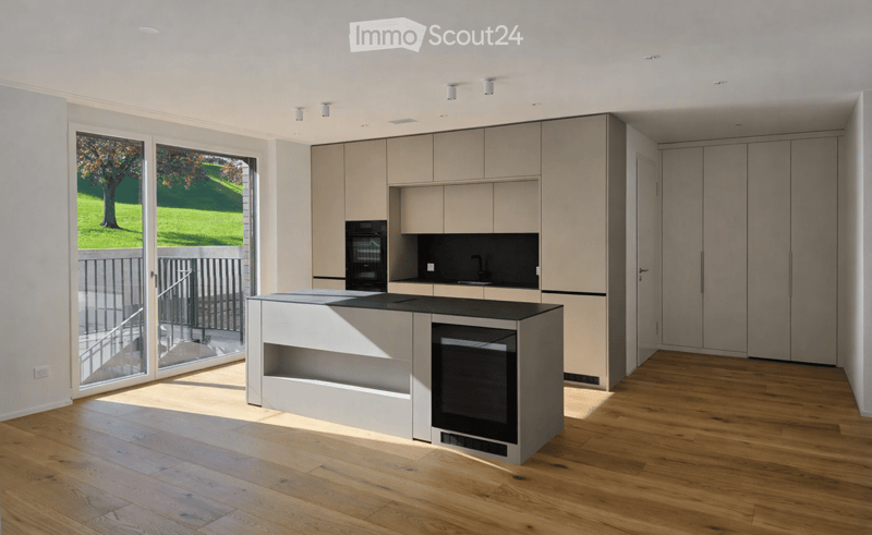 Modern kitchen with light wood cabinets, a central island, and built-in appliances, featuring large windows and a view of a green outdoor area.