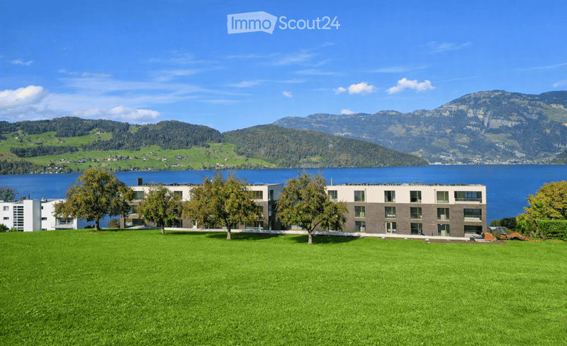 Modern multi-story building with a combination of white and brown facades, surrounded by a well-maintained green lawn and trees, with a scenic lake and mountain backdrop.